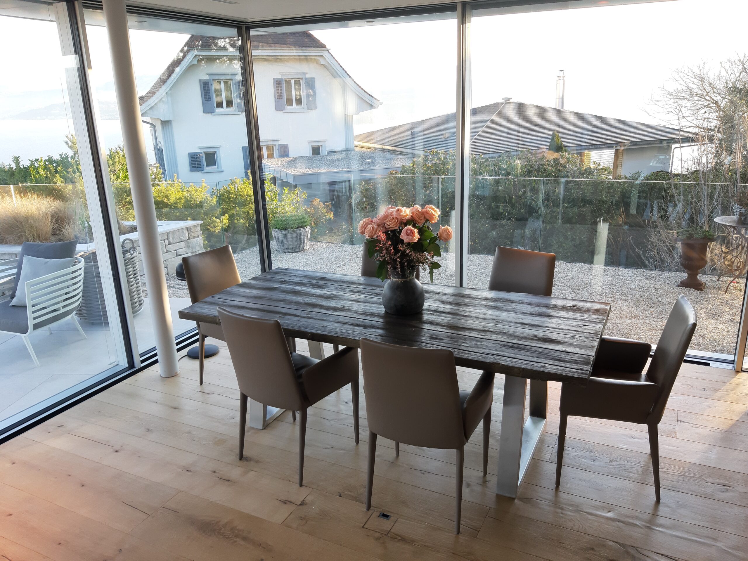 Esstisch aus Altholz mit sechs Stühlen vor grossen Fensterfronten